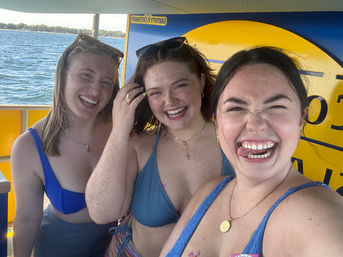 Three friends in swimsuits taking a playful selfie on a bright yellow pedal boat at a sunny lake with water and shoreline in the background