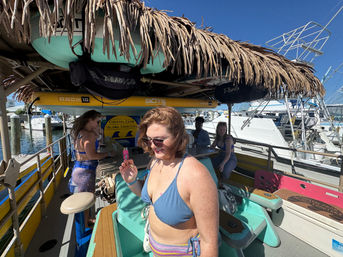 Women in swimsuits enjoying a tiki-canopied pontoon boat at a coastal marina, paddleboards stowed overhead and boats docked nearby under a clear blue sky.
