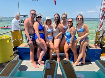 Seven friends in colorful swimsuits smiling and posing on a party boat over turquoise coastal waters on a sunny day