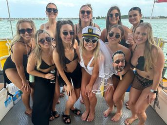 Smiling group of women in swimsuits on a party boat over turquoise coastal water, bride-to-be wearing a captain's hat and veil holding a playful face cutout