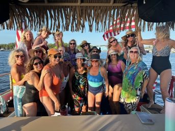 Smiling group of women in colorful swimsuits and sun hats on a sunny boat party under a thatched tiki roof with an American flag and marina waterfront in the background.