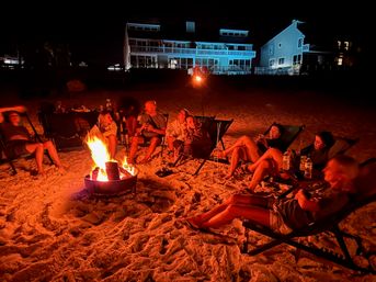 Group relaxing around a glowing beach bonfire at night, lounging in folding chairs on the sandy shore with illuminated beachfront houses in the background.