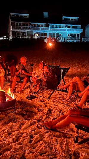 Group relaxing around a glowing beach bonfire at night, lounging in folding chairs on the sandy shore with illuminated beachfront houses in the background.