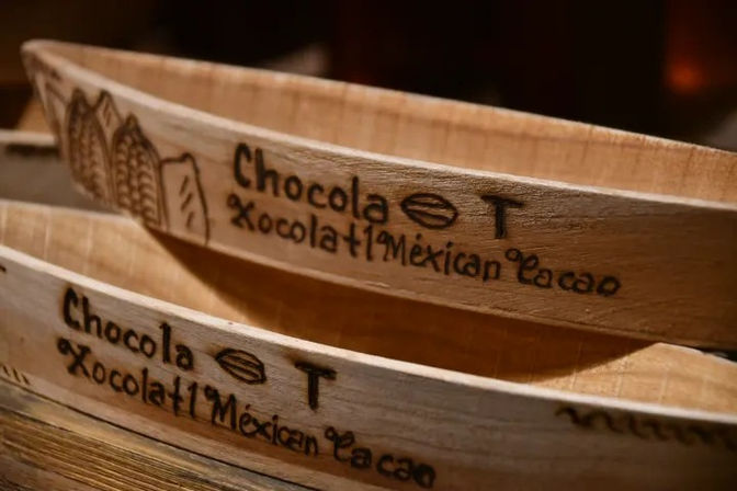 Close-up of rustic wooden trays engraved with 'Chocola Xocolatl Mexican cacao' and a small cacao bean sketch, evoking Mexican chocolate and artisanal craft.
