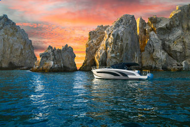 Sleek motorboat floating beside dramatic rocky sea cliffs at sunset, fiery sky reflected on turquoise ocean water.
