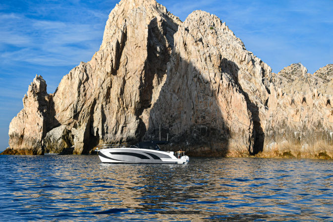 Sleek white motorboat bobbing beside towering sunlit coastal rock cliffs rising from deep blue sea under a clear sky