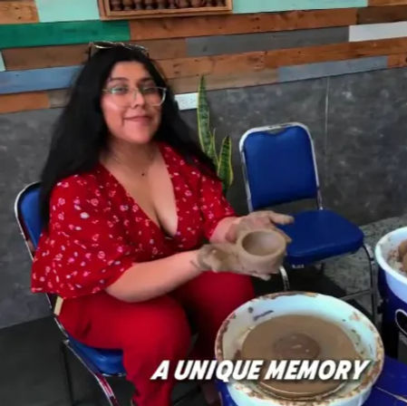 Person in a red outfit smiling while shaping a clay bowl on a pottery wheel in a ceramics studio with blue chairs and a wooden accent wall, text overlay reads “A UNIQUE MEMORY”.