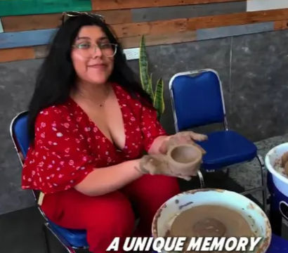 Person in a red outfit smiling while shaping a clay bowl on a pottery wheel in a ceramics studio with blue chairs and a wooden accent wall, text overlay reads “A UNIQUE MEMORY”.