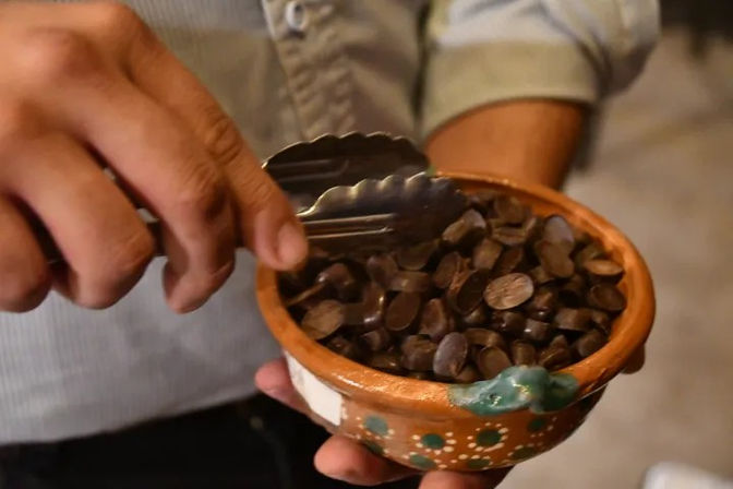 Close-up of hands using metal tongs to pick dark chocolate discs from a small hand-painted terracotta bowl with dotted decoration.