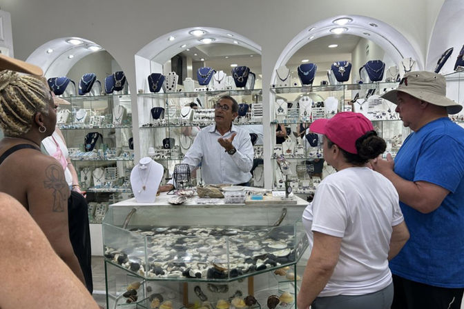 Shopkeeper animatedly explaining pieces to a small group of shoppers inside a bright jewelry boutique, glass cases filled with necklaces, rings and a shell-filled display counter.