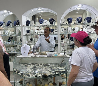 Shopkeeper animatedly explaining pieces to a small group of shoppers inside a bright jewelry boutique, glass cases filled with necklaces, rings and a shell-filled display counter.