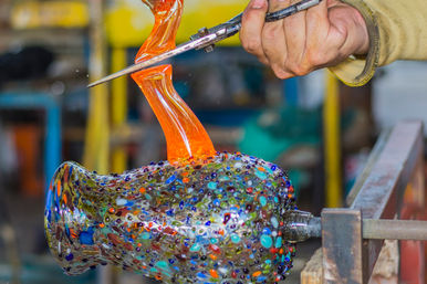 Close-up of a glassblower shaping a colorful speckled blown-glass vase with glowing orange molten glass and metal shears in a studio