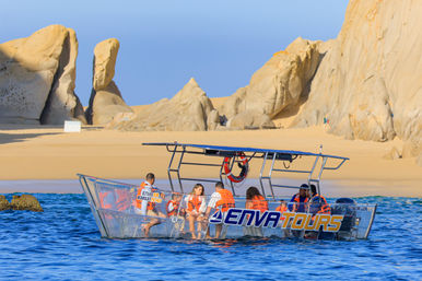 Glass-sided tour boat with passengers in orange life jackets cruising bright blue water off a sunny, sandy desert coastline framed by dramatic sandstone rock formations.