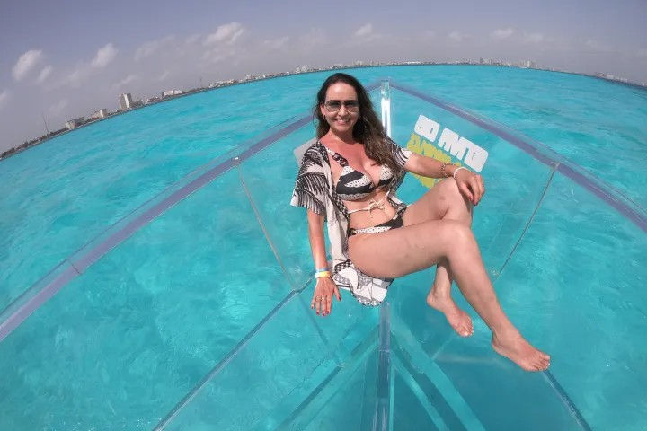 Smiling woman in a patterned bikini and sunglasses lounging on a clear-hulled boat above vivid turquoise tropical water, with a sunny sky and a distant coastal skyline