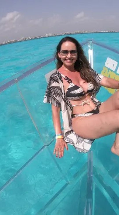 Smiling woman in a patterned bikini and sunglasses lounging on a clear-hulled boat above vivid turquoise tropical water, with a sunny sky and a distant coastal skyline