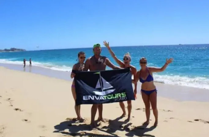 Four people in swimsuits on a sunny sandy beach by a turquoise ocean, holding a black banner with a sailboat logo and waving, with footprints in the sand and two distant beachgoers near the shoreline.