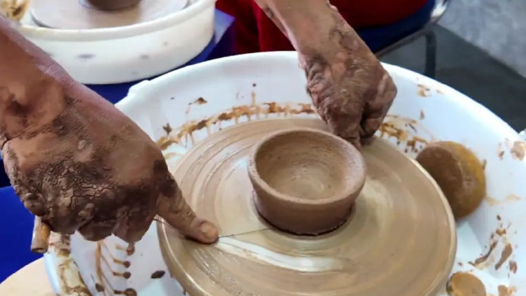 Close-up of clay-covered hands shaping a small handmade ceramic bowl on a spinning pottery wheel in a studio, with sponge and clay splatters.