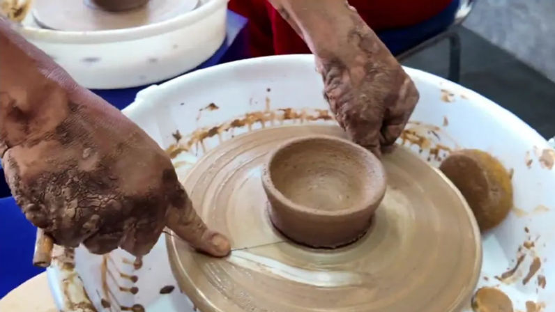 Close-up of clay-covered hands shaping a small handmade ceramic bowl on a spinning pottery wheel in a studio, with sponge and clay splatters.