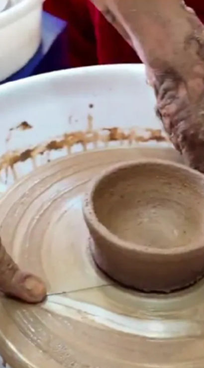 Close-up of clay-covered hands shaping a small handmade ceramic bowl on a spinning pottery wheel in a studio, with sponge and clay splatters.