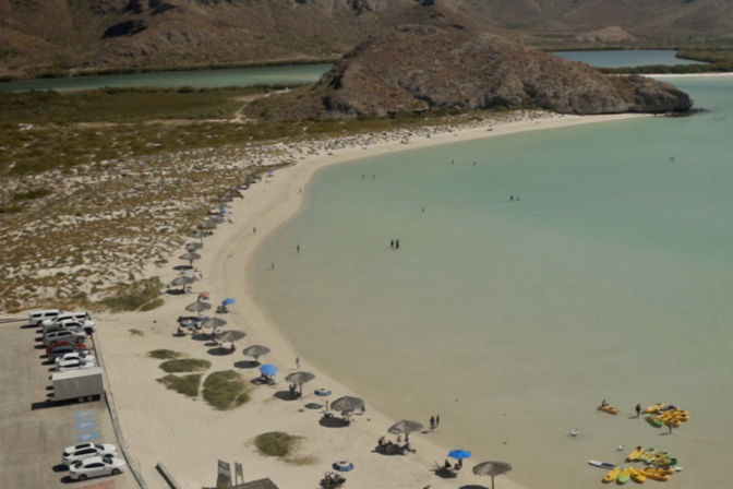 Aerial view of a crescent-shaped turquoise lagoon and white-sand beach dotted with palapa umbrellas, scattered beachgoers, kayaks on the shore and a small parking area, with rocky hills in the background.