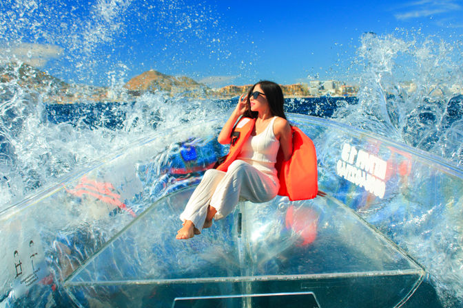Woman in sunglasses wearing an orange life jacket and white outfit seated in a transparent kayak as ocean waves splash dramatically under a bright blue sky with a rocky coastal backdrop