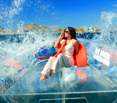 Woman in sunglasses wearing an orange life jacket and white outfit seated in a transparent kayak as ocean waves splash dramatically under a bright blue sky with a rocky coastal backdrop