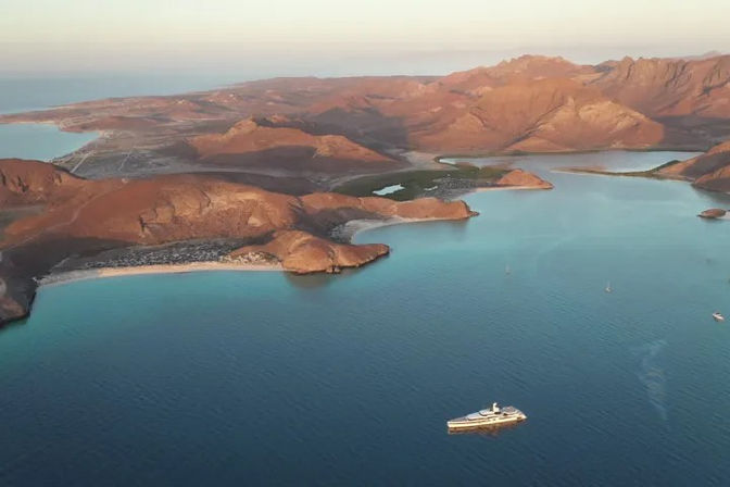Aerial view of a turquoise coastal bay and arid rocky islands with a small yacht anchored in calm blue water and golden mountains lit by sunset.