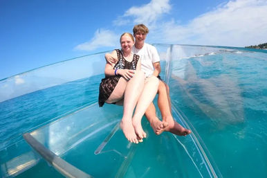 Two smiling people sitting on the transparent bow of a glass-bottom boat over turquoise tropical ocean under a bright blue sky