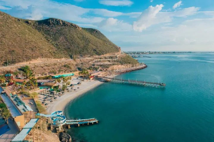 Aerial view of a sunny curved sandy beach and long wooden pier on a turquoise bay, backed by green hills, winding coastal road, beach umbrellas and a waterslide.