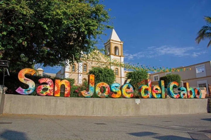 Vibrant oversized 'San José del Cabo' letters in a sunny plaza in San José del Cabo, Mexico, with a historic white church tower, palm tree, and bright blue sky