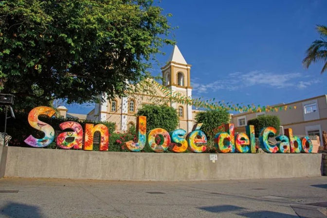 Vibrant oversized 'San José del Cabo' letters in a sunny plaza in San José del Cabo, Mexico, with a historic white church tower, palm tree, and bright blue sky