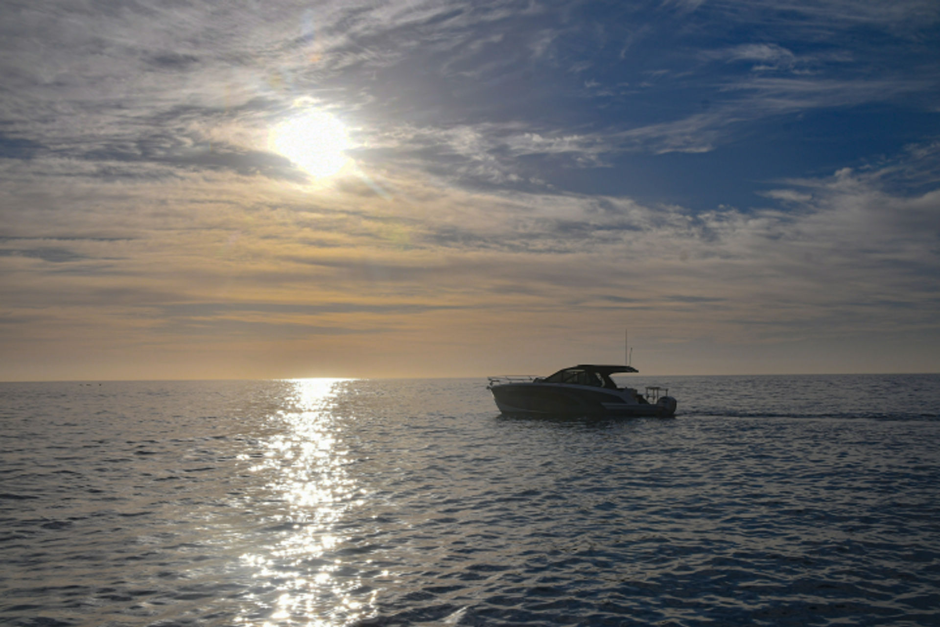 Serene motorboat on calm open sea at sunset, sun reflecting in a glittering water path beneath streaky clouds.