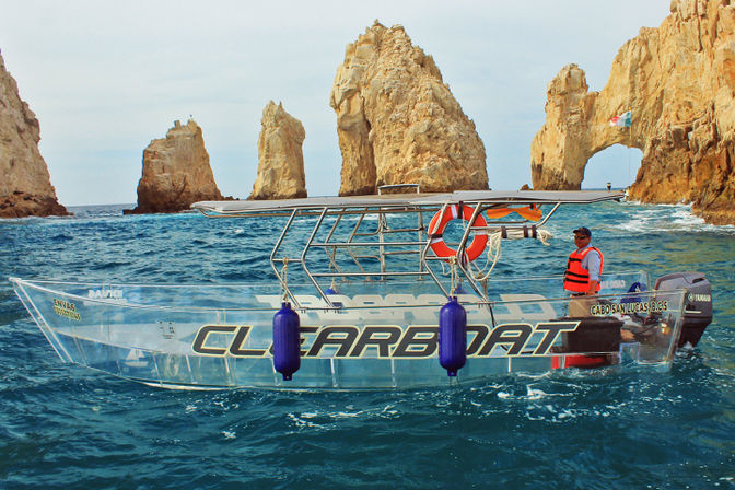 Clear-hull tour boat with bright fenders and life ring, crew member in an orange life vest cruising turquoise waters by dramatic sea rock arches at Cabo San Lucas, Mexico