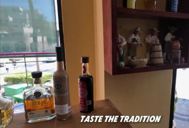 Three bottles of Mexican spirits on a wooden bar counter by a sunlit window overlooking a seaside street, a shelf with folkloric figurines, and the text overlay "TASTE THE TRADITION".