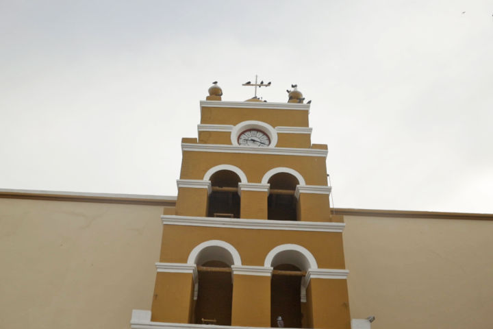 Historic yellow colonial-style clock and bell tower with white arched openings and pigeons perched on top against an overcast sky