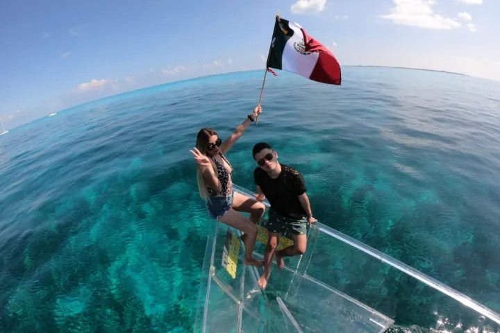 Two people perched on the glass bow of a clear-bottom boat over turquoise Mexican waters, one holding a waving Mexican flag with coral visible below