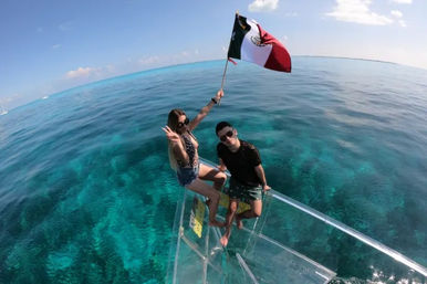 Two people perched on the glass bow of a clear-bottom boat over turquoise Mexican waters, one holding a waving Mexican flag with coral visible below