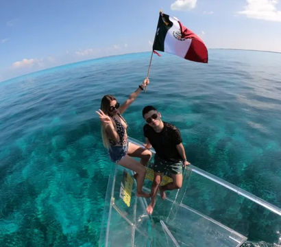 Two people perched on the glass bow of a clear-bottom boat over turquoise Mexican waters, one holding a waving Mexican flag with coral visible below