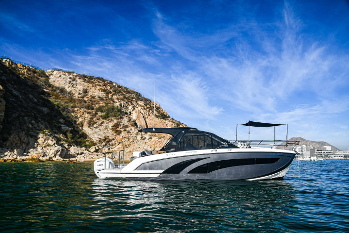 Sleek gray-and-white cabin cruiser anchored in a calm turquoise bay near a rocky coastal cove under a bright blue sky with a marina visible on the distant shore