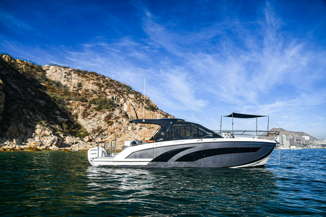 Sleek gray-and-white cabin cruiser anchored in a calm turquoise bay near a rocky coastal cove under a bright blue sky with a marina visible on the distant shore