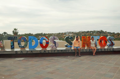 Bright painted 'Todos Santos' public art sign on a tiled plaza in Baja California Sur, Mexico, colorful letters with two visitors seated on the ledge, palm trees and hills in the background