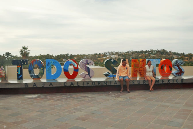 Bright painted 'Todos Santos' public art sign on a tiled plaza in Baja California Sur, Mexico, colorful letters with two visitors seated on the ledge, palm trees and hills in the background