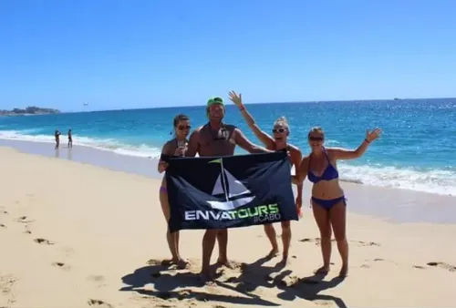 Four people in swimsuits cheer while holding a tour banner on a sunny Cabo San Lucas beach with turquoise water and a clear blue sky.