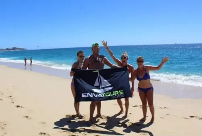 Four people in swimsuits cheer while holding a tour banner on a sunny Cabo San Lucas beach with turquoise water and a clear blue sky.