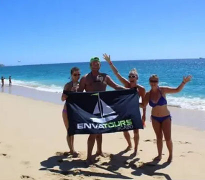Four people in swimsuits cheer while holding a tour banner on a sunny Cabo San Lucas beach with turquoise water and a clear blue sky.