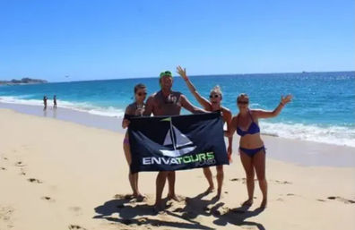 Four adults in swimwear holding a black tour banner and waving on a sunny sandy Cabo beach with turquoise ocean and clear blue sky.