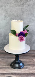 Elegant two-tier white buttercream cake decorated with pink roses, purple blooms and green leaves on a black pedestal cake stand, set on a wooden tabletop against a gray textured backdrop