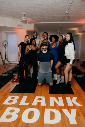 Energetic group of people in workout clothes posing on yoga mats in a bright indoor fitness studio, some holding small orange dumbbells, wood floor with bold white lettering and warm studio lights.