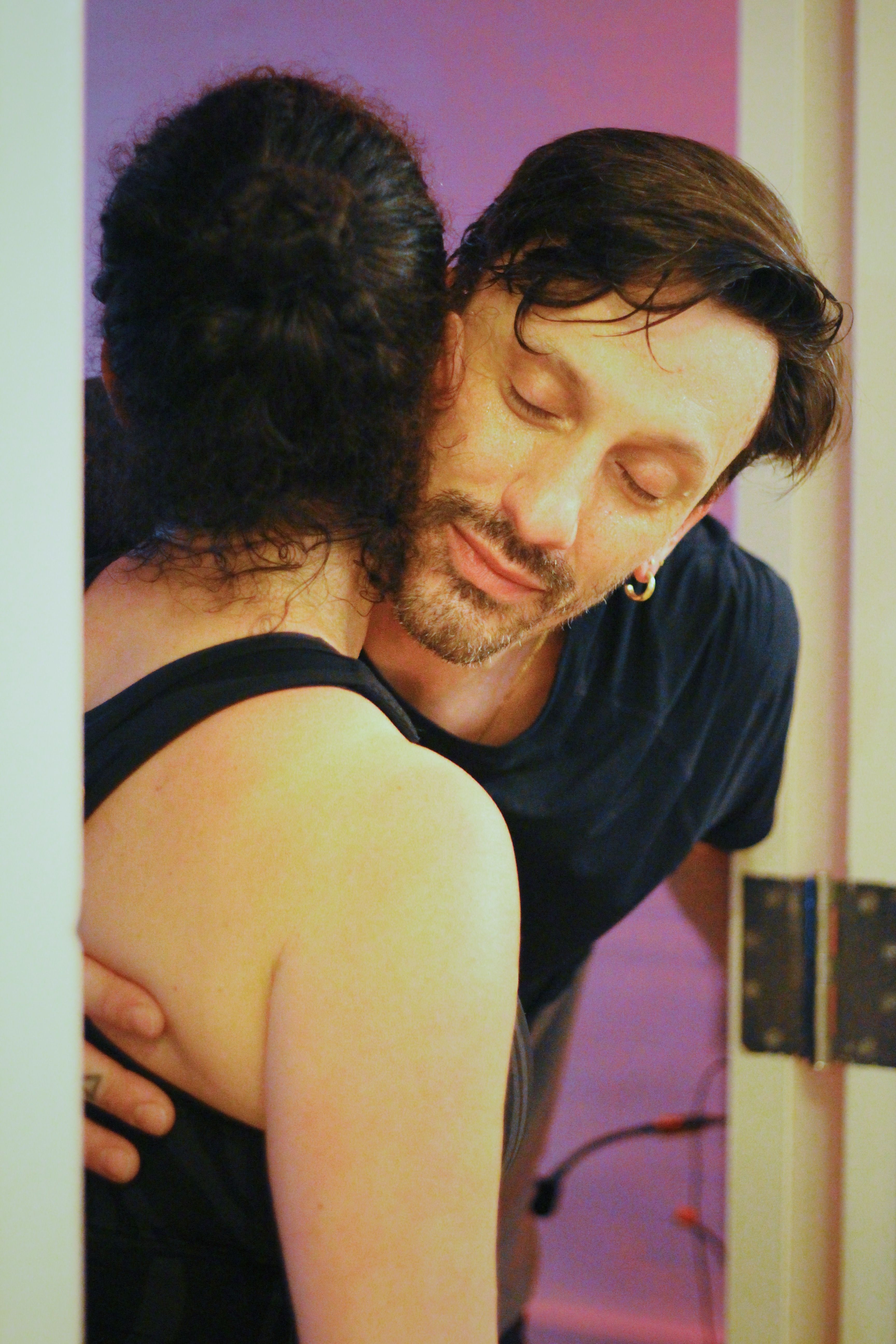 Couple sharing a tender hug in a doorway, man with beard and earring resting his head on partner’s shoulder under warm purple indoor lighting