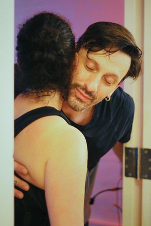 Couple sharing a tender hug in a doorway, man with beard and earring resting his head on partner’s shoulder under warm purple indoor lighting